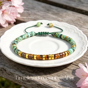 May include: A handmade bracelet featuring turquoise, brown, and gold beads. The bracelet is strung on a green cord with adjustable ends. The bracelet is displayed on a white plate with the text "Timeless Treasures Aha" and the website address.