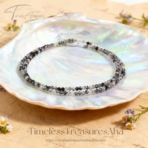 May include: A delicate necklace featuring small, round beads in shades of black, white, and clear, strung together. The necklace is displayed on a white, iridescent seashell. The text "Timeless Treasures" and the Etsy shop URL are visible.