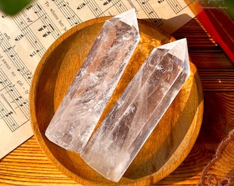 May include: Two clear quartz crystal points, also known as double terminated crystals, are displayed on a wooden bowl. The crystals are long and slender, with a pointed tip at each end. The crystals are clear and translucent, with a slight shimmer.