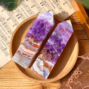 May include: Two amethyst crystal obelisks with purple and white banding, displayed on a wooden plate with sheet music in the background. The crystals have a polished, pointed top and a layered, natural appearance.