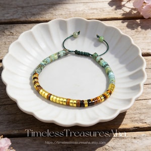 May include: A beaded bracelet with turquoise, gold, and brown accents. The bracelet is displayed on a white, scalloped plate. The text "Timeless Treasures Aha" and a website address are visible at the bottom.