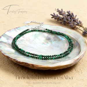 May include: A delicate, layered necklace featuring small, faceted emerald green beads. The necklace is displayed on a pearlescent, iridescent shell-shaped dish, with a sprig of lavender in the background. The words "Timeless Treasures" are visible.