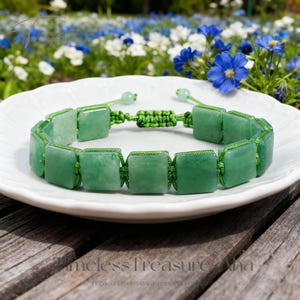 May include: A green aventurine bracelet with square beads and a macrame closure. The bracelet is displayed on a white plate against a backdrop of flowers and a wooden surface.