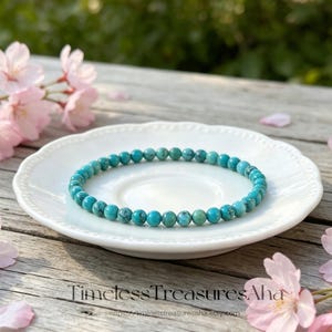 May include: A turquoise beaded bracelet rests on a white, scalloped-edge plate. The bracelet is composed of small, round, turquoise beads. Pink cherry blossoms and the text "Timeless Treasures Aha" are in the background.