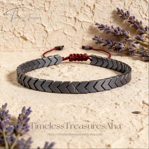 Puede incluir: Una pulsera de hematita gris oscuro con una serie de cuentas en forma de cheurón dispuestas en una banda continua. La pulsera incluye una cuenta central grabada con la letra Y y se asegura con un mecanismo de cierre de cordón rojo ajustable.