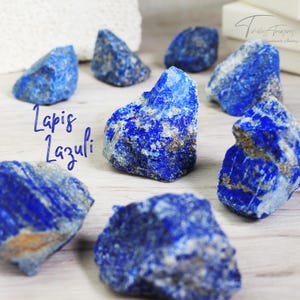May include: A collection of eight rough, natural Lapis Lazuli stones. The stones are a deep blue color with white and gold flecks. The stones are arranged on a light wood surface. The text "Lapis Lazuli" is written in white on the surface.