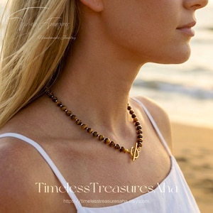 May include: A beaded necklace with tiger eye stones and a gold-colored clasp. The necklace is worn by a person wearing a white top. The image includes the text "Timeless Treasures Aha" and "Handmade Jewelry".