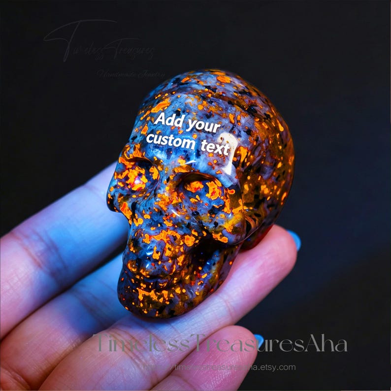 Puede incluir: Una calavera de piedra pulida y tallada con un patr&oacute;n moteado de azul profundo, negro y motas de color naranja vibrante. La zona de la frente muestra el texto blanco "Add your custom text" en una fuente sans-serif sobre la superficie oscura.