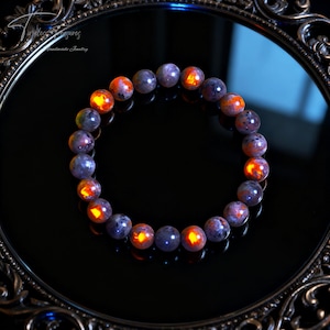 May include: A beaded bracelet featuring round, polished stones in shades of purple and orange. The stones have a glowing, fiery appearance. The bracelet is displayed on a reflective black surface, with a decorative frame in the background. The jewelry is handmade.
