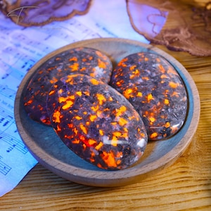 May include: Three oval-shaped stones with a dark gray base and vibrant orange and yellow inclusions are displayed in a wooden bowl. The stones have a polished surface. The bowl sits on a wooden surface.