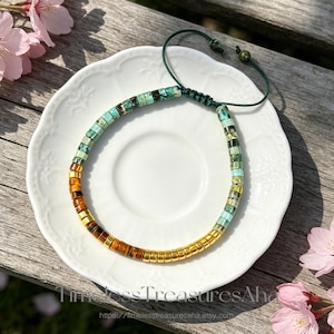 May include: A beaded bracelet featuring a mix of turquoise-colored stone discs, brown tiger eye beads, and metallic gold-toned cylindrical beads. The bracelet includes a dark green adjustable cord closure with two small matching green beads at the ends.