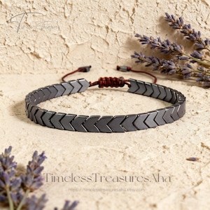 May include: A dark gray hematite bracelet with an arrow design. The bracelet is on a brown cord with an adjustable knot closure. The background is a textured beige surface with sprigs of lavender. The text "Timeless Treasures Aha" is at the bottom.