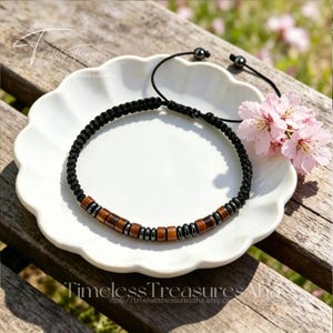 Puede incluir: Una pulsera trenzada negra con cuentas de madera y metálicas. La pulsera se exhibe en un plato blanco con borde festoneado, con flores rosas y el texto "Timeless Treasures" en el fondo.