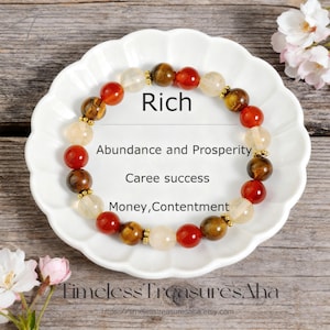 May include: A beaded bracelet with red, brown, yellow, and clear beads, displayed on a white plate. The word "Rich" is written above the bracelet, with additional text below. The bracelet is from Timeless Treasures Aha.