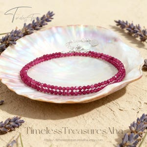 May include: A double-strand, faceted ruby gemstone bracelet with a silver clasp, resting in a white shell. The bracelet is a vibrant shade of red. The shell is on a light brown surface, with lavender sprigs around it. The text "Timeless Treasures Aha" is at the bottom.