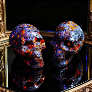 Puede incluir: Dos esculturas de calaveras talladas en piedra de color morado oscuro y negro con inclusiones de color naranja y rojo vibrante en toda la superficie. Las calaveras presentan cuencas oculares, cavidades nasales y dientes detallados, reflej&aacute;ndose en una superficie oscura.