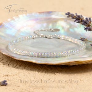 Puede incluir: Una delicada pulsera de cuentas iridiscentes con un cierre plateado descansa sobre una concha nacarada. La pulsera está compuesta por pequeñas cuentas redondas que brillan con un efecto arcoíris. La imagen incluye el texto "Timeless Treasures" y una dirección web.