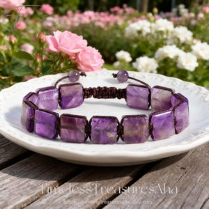 May include: A square amethyst bracelet with a macrame cord. The bracelet is displayed on a white plate with a floral background. The bracelet is made of square amethyst stones and a brown macrame cord.