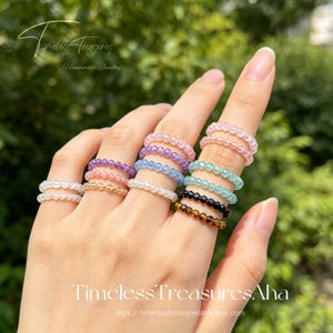 May include: A collection of elastic rings made from small, faceted, multi-colored gemstone beads. The rings feature various colors including light pink, purple, blue, white, black, and brown, worn stacked across multiple fingers.