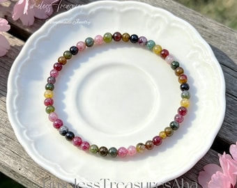 May include: A beaded bracelet featuring a variety of colorful tourmaline gemstones. The bracelet is displayed on a white plate with a scalloped edge, with the words "Timeless Treasures" and "Handmade Jewelry" visible in the background.