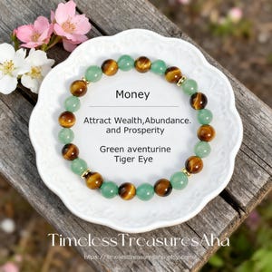 May include: A beaded bracelet featuring alternating green aventurine and tiger eye stones. The bracelet is displayed on a white, scalloped plate with text that reads "Money" and "Attract Wealth, Abundance, and Prosperity."