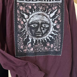 May include: A maroon long-sleeved button-up shirt with a black and white patch on the back. The patch features a sun with a face and flowers, and the word "SUBlime" in white letters.