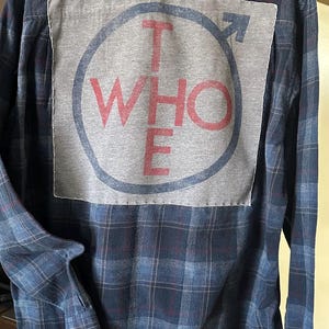 May include: A blue plaid flannel shirt with a gray patch sewn on the back. The patch features a circle with the word "WHO" in red letters and an arrow pointing to the right.