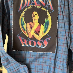 May include: Blue plaid button-down shirt with a large black patch featuring the text "DIANA ROSS" and an illustration of Diana Ross. The shirt has long sleeves and a classic collar.