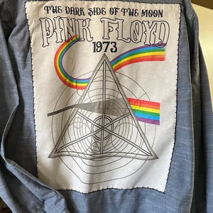 May include: A blue denim button-down shirt with a white patch on the back. The patch features a rainbow, a triangle, and the text "The Dark Side of the Moon Pink Floyd 1973".