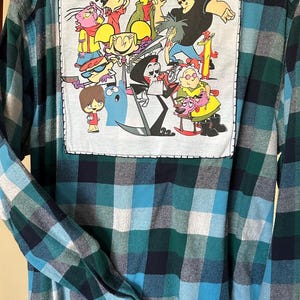 May include: A blue and green plaid flannel shirt with a cartoon character patch on the back. The patch features various cartoon characters from Cartoon Network, including Dexter's Laboratory, Ed, Edd n Eddy, Johnny Bravo, Courage the Cowardly Dog, and Powerpuff Girls. The patch also includes the Cartoon Network logo.