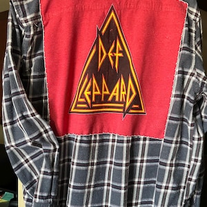 May include: A blue and white plaid flannel shirt with a red patch on the back. The patch features a black triangle with the words "Def Leppard" in yellow lettering.