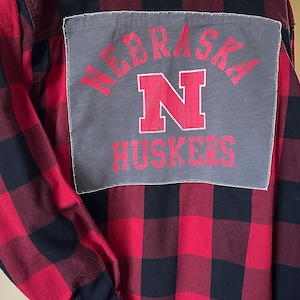 May include: Red and black plaid flannel shirt with a gray patch on the back. The patch features the word "NEBRASKA" arched above a large red "N" and the word "HUSKERS" below. The shirt has long sleeves and a button-down front.