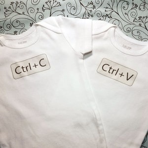 May include: Two white baby onesies with brown text. The onesie on the left says "Ctrl+C" and the onesie on the right says "Ctrl+V".