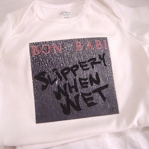 May include: White baby onesie with a black and white graphic of a record album cover. The album title is "BON BABI" and the album artist is "SLIPPERY WHEN WET".