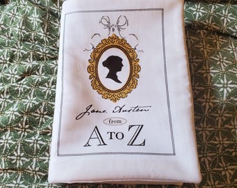 Peut inclure: Sac fourre-tout blanc avec une silhouette dans un cadre doré, un nœud et le texte "Jane Austen from A to Z". Le sac est rectangulaire et en tissu. Il repose sur un tissu vert à motifs.