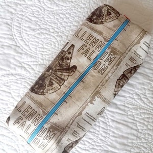 Puede incluir: Una bolsa de cremallera beige con una cremallera azul. La bolsa tiene un estampado marrón y blanco del Millennium Falcon y el texto "Millennium Falcon".