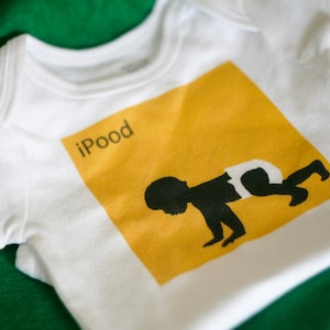 Pode incluir: Um macacão branco para bebê com um quadrado amarelo que apresenta uma silhueta preta de um bebê engatinhando. O quadrado tem o texto "iPod" em preto.
