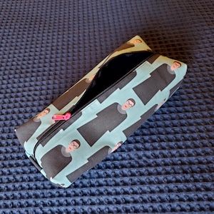 May include: A blue and black zippered pencil case with a repeating pattern of a woman wearing a black robe and glasses. The zipper pull is pink.