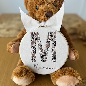 May include: A brown plush teddy bear holding a circular wooden embroidery hoop. The hoop contains off-white fabric featuring a large letter M embroidered with brown, white, and black floral patterns, with the name Marceau stitched below.