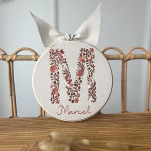 May include: A circular embroidered wall hanging with the letter "M" in a floral pattern and the name "Marcel" below. The embroidery uses brown and white threads on a beige background, with a beige bow at the top.