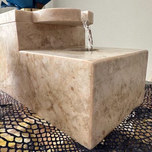 Pu&ograve; includere: Una fontana in pietra beige con una cascata d'acqua. La fontana ha una base rettangolare e una parte superiore rettangolare pi&ugrave; piccola con un becco curvo. L'acqua scorre dal becco nella vasca inferiore.
