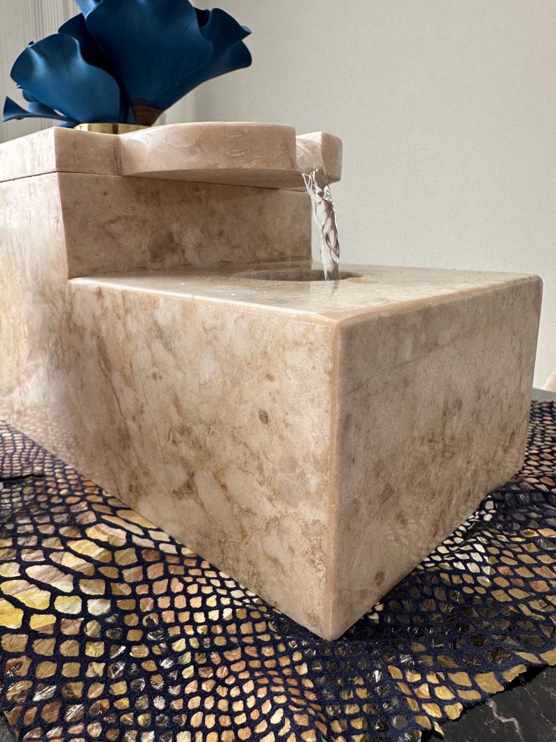Pu&ograve; includere: Una fontana in pietra beige con una cascata d'acqua. La fontana ha una base rettangolare e una parte superiore rettangolare pi&ugrave; piccola con un becco curvo. L'acqua scorre dal becco nella vasca inferiore.
