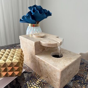 Pu&ograve; includere: Una fontana da tavolo in pietra beige con una cascata d'acqua. La fontana &egrave; rettangolare e ha una superficie liscia e lucida. &Egrave; appoggiata su una superficie con motivo nero e dorato.