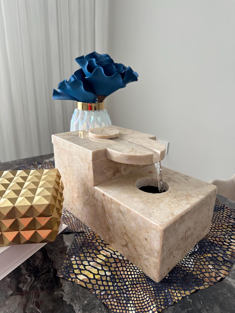Pu&ograve; includere: Una fontana da tavolo in pietra beige con una cascata d'acqua. La fontana &egrave; rettangolare e ha una superficie liscia e lucida. &Egrave; appoggiata su una superficie con motivo nero e dorato.