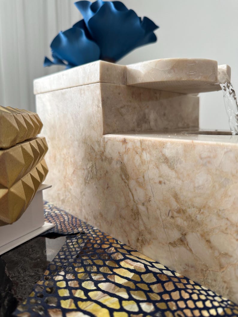 Pu&ograve; includere: Una fontana in marmo bianco con una cascata d'acqua. La fontana ha una base rettangolare e un design a pi&ugrave; livelli. Il livello superiore ha un beccuccio curvo che dirige il flusso d'acqua. La fontana &egrave; decorata con un fiore blu.