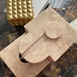 Pu&ograve; includere: Una fontana da tavolo in marmo beige con un'apertura circolare nella parte superiore e una base rettangolare. La fontana ha una superficie liscia e lucida e un design moderno.