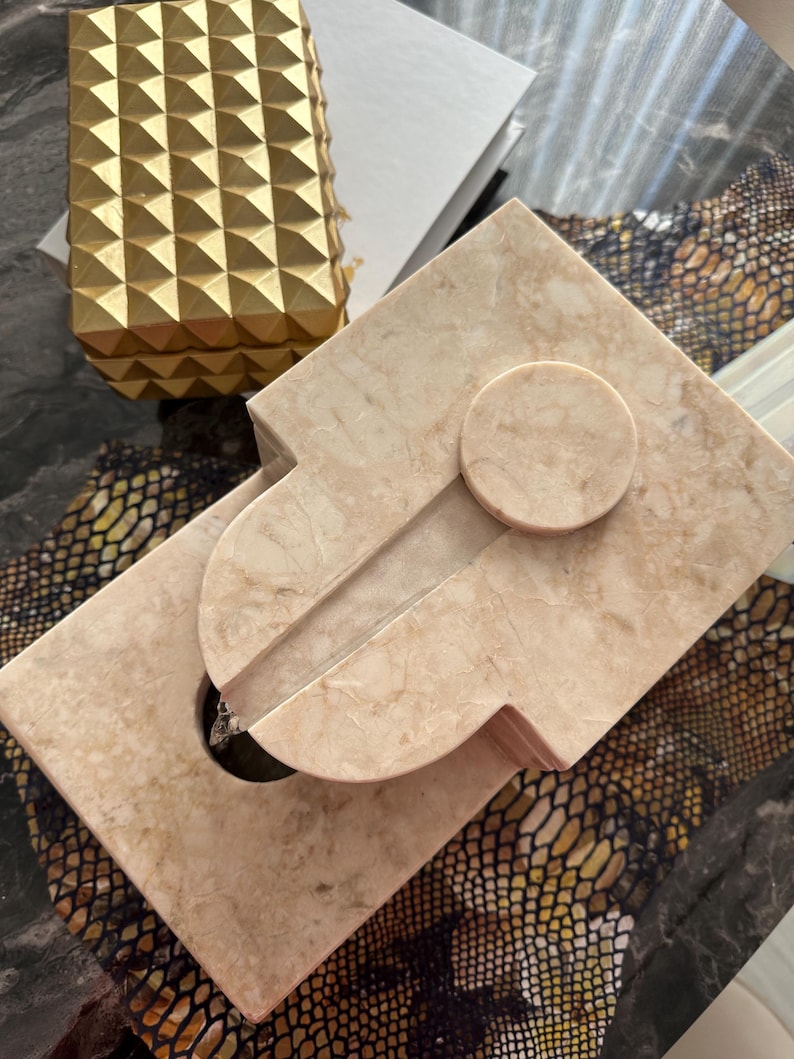 Pu&ograve; includere: Una fontana da tavolo in marmo beige con un'apertura circolare nella parte superiore e una base rettangolare. La fontana ha una superficie liscia e lucida e un design moderno.