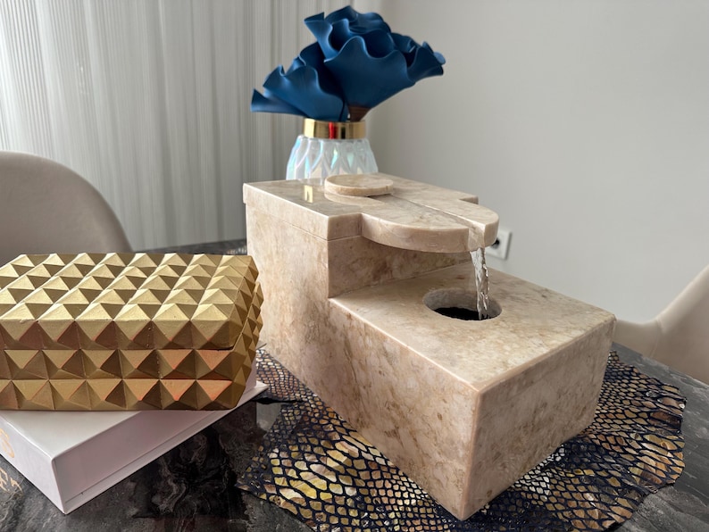 Pu&ograve; includere: Una fontana da tavolo in pietra beige con una cascata d'acqua. La fontana &egrave; su un tavolo con una scatola decorativa dorata e una superficie con motivo.
