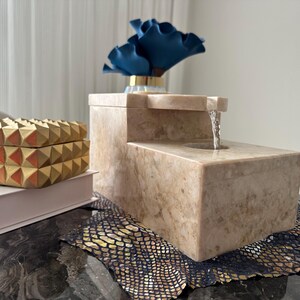 Pu&ograve; includere: Una fontana da tavolo in pietra beige con una cascata d'acqua. La fontana &egrave; su una superficie nera con un tappetino a motivi dorati e neri.