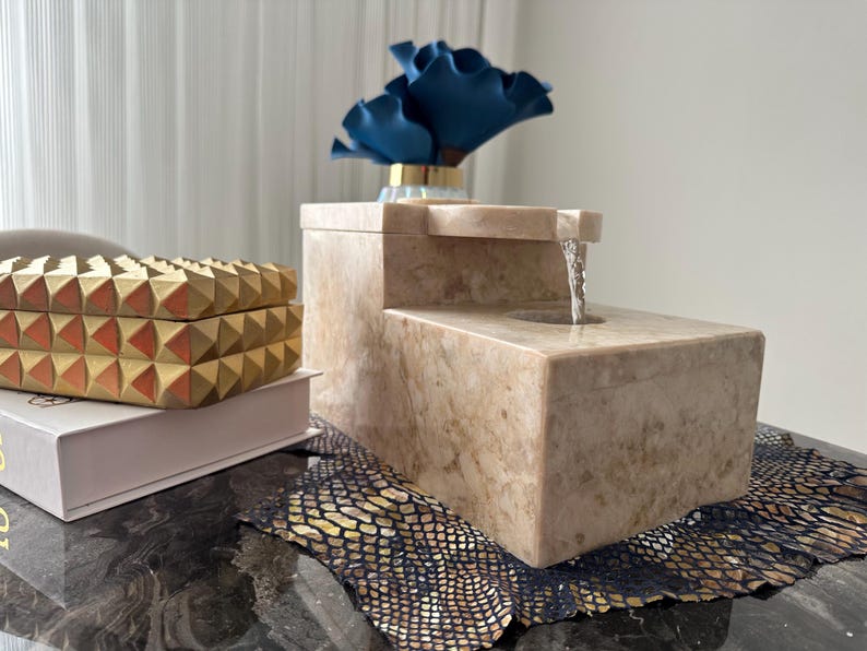 Pu&ograve; includere: Una fontana da tavolo in pietra beige con una cascata d'acqua. La fontana &egrave; su una superficie nera con un tappetino a motivi dorati e neri.